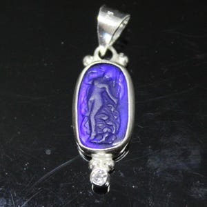 May include: A silver pendant with a deep blue oval-shaped center featuring a carved figure. The pendant has a small, clear gemstone at the bottom and a loop at the top for a chain. The figure is a classical depiction of a woman.