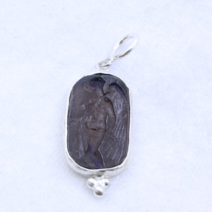 May include: A silver pendant with a dark purple, rectangular stone. The stone features a carved image of a winged figure. The pendant has a loop at the top and a decorative cluster of three small spheres at the bottom.