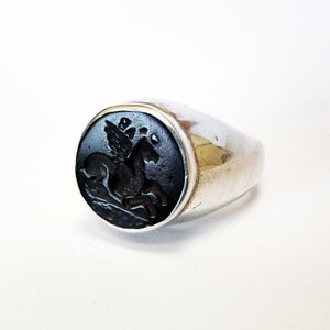 May include: A silver ring with a black onyx stone featuring a carved image of a winged horse.