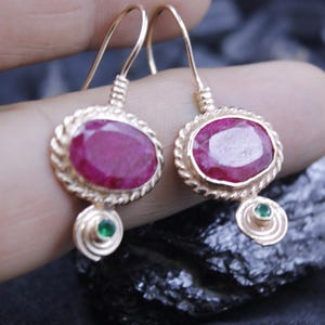 May include: A pair of gold-colored drop earrings. Each earring features an oval, deep red gemstone set within a twisted gold frame. A small green gemstone is suspended below each oval stone, adding a pop of color.