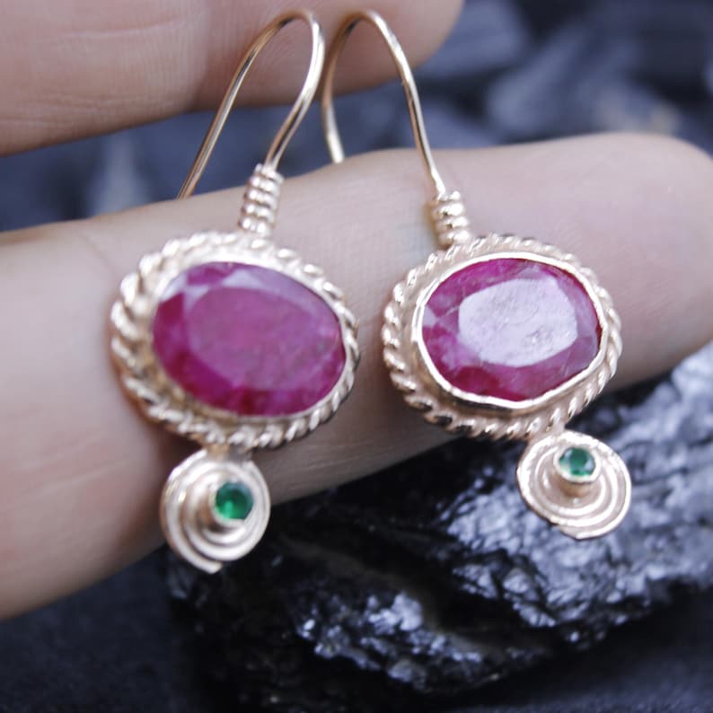 May include: A pair of gold-colored drop earrings. Each earring features an oval, deep red gemstone set within a twisted gold frame. A small green gemstone is suspended below each oval stone, adding a pop of color.