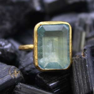 Peut inclure: Une bague en or ornée d'une grande pierre précieuse rectangulaire vert clair. La pierre est sertie dans une lunette en or et repose sur un anneau en or. La bague est exposée sur un lit de pierres sombres et rugueuses.