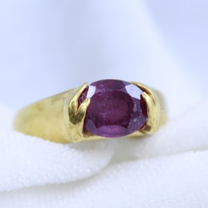 May include: A gold ring with a large, oval-shaped purple gemstone set in the center. The ring band is thick and has a unique design.