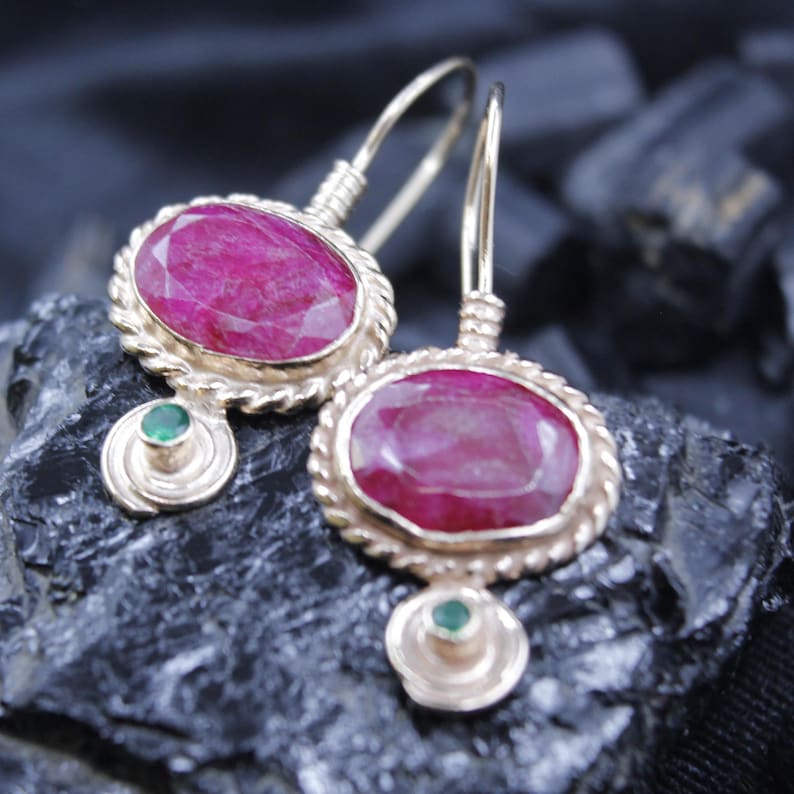 May include: Gold-toned earrings featuring large, oval-cut ruby gemstones. The rubies are set in a textured, rope-like border. Each earring has a small, round emerald accent below the ruby, and a wire hook for pierced ears.