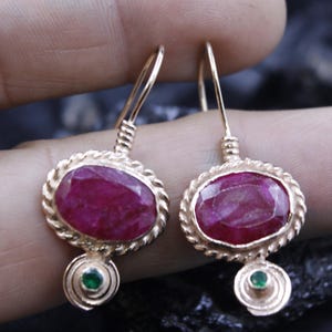 May include: Gold-toned dangle earrings featuring oval, deep red gemstones. Each earring has a twisted rope-like border around the main stone and a small green gemstone at the bottom. The earrings are designed with a hook closure.