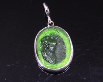Puede incluir: Un colgante verde con un marco plateado. El colgante presenta un relieve de una figura desnuda dentro de una forma ovalada. La figura es de color verde oscuro y el fondo es verde claro. El colgante tiene un pequeño lazo en la parte superior para una cadena.