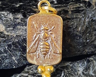 May include: A gold-colored pendant with a rectangular face featuring a detailed bee design. The pendant has a small loop at the top for a chain and a cluster of three small spheres at the bottom. The background is a dark, textured surface.