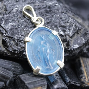 May include: A silver pendant with a light blue oval-shaped center featuring a religious figure. The pendant has a loop at the top for a chain and is set against a dark, textured background.