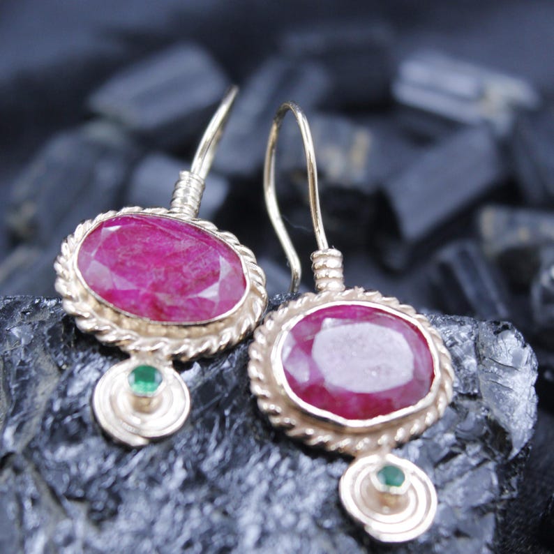 May include: A pair of gold-colored earrings with oval, deep red gemstones. The gemstones are set in a twisted rope-like frame. Each earring has a small, round green gemstone below the oval stone.