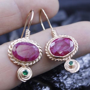 May include: A pair of gold-colored earrings featuring oval-shaped, deep red gemstones. Each earring has a smaller green gemstone at the bottom and a twisted rope-like border around the main stone. The earrings are displayed against a dark background.