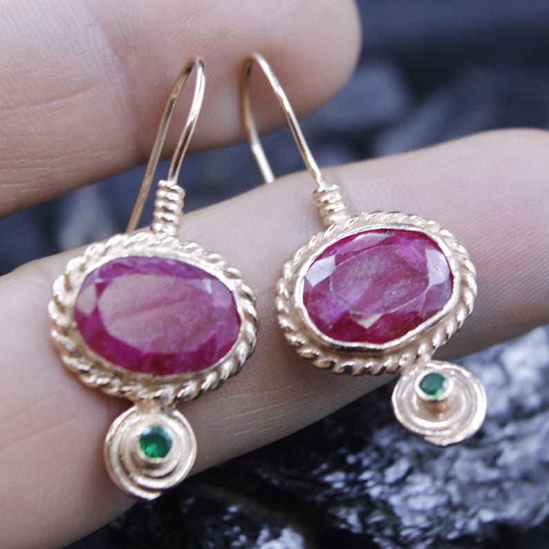 May include: A pair of gold-colored earrings featuring oval-shaped, deep red gemstones. Each earring has a smaller green gemstone at the bottom and a twisted rope-like border around the main stone. The earrings are displayed against a dark background.