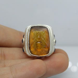 May include: A silver ring with a rectangular amber stone featuring an engraved bee design. The ring is accented with small blue stones.