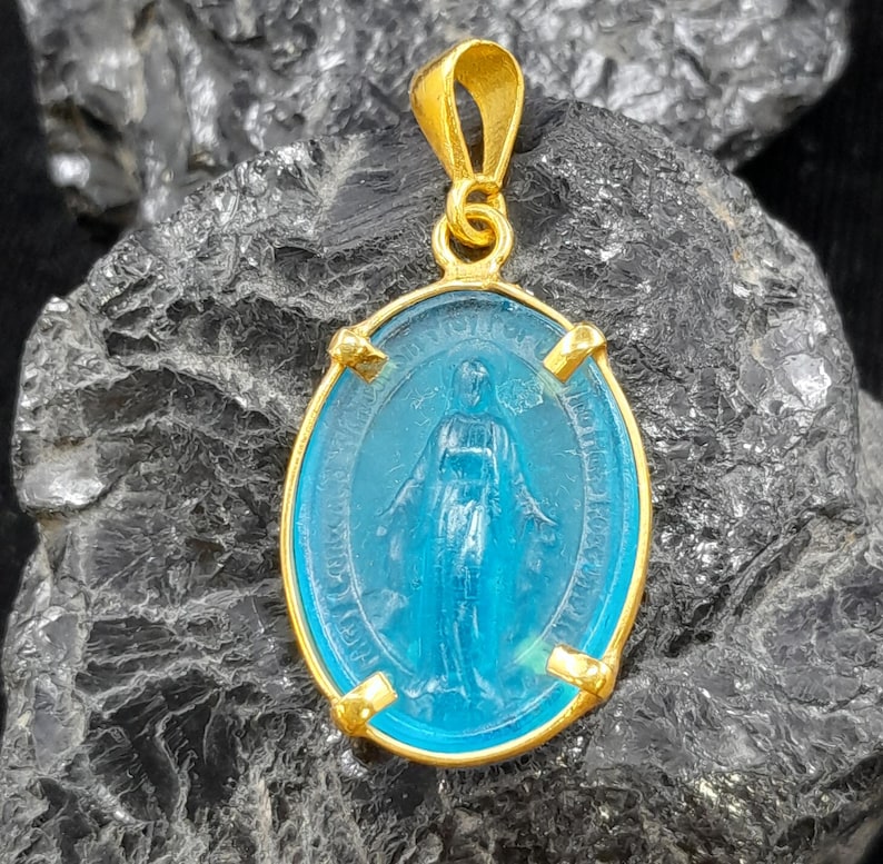 Puede incluir: Pendiente de oro con un camafeo de vidrio azul de la Virgen Mar&iacute;a. El camafeo tiene forma ovalada y tiene un marco de oro con cuatro acentos decorativos.