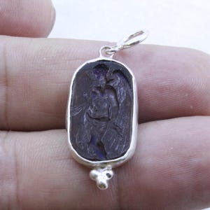 May include: A silver pendant with a dark purple, oval-shaped stone featuring an engraved image of an angel with wings. The pendant has a small loop at the top and a decorative cluster at the bottom.