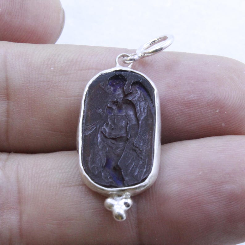 May include: A silver pendant with a dark purple, oval-shaped stone featuring an engraved image of an angel with wings. The pendant has a small loop at the top and a decorative cluster at the bottom.