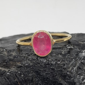 May include: A gold ring featuring a large, oval-cut, bright pink gemstone. The ring's band is thin and gold-colored, complementing the vibrant hue of the gemstone. The ring is set against a dark, textured background.