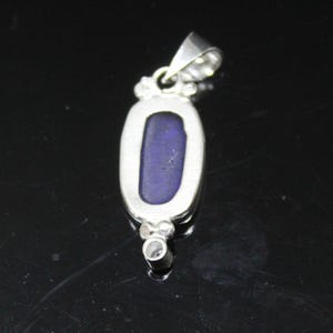 May include: A silver pendant with an oval, deep blue stone. The pendant has a bail at the top for a chain and a small decorative element at the bottom. The stone is set in a silver bezel.