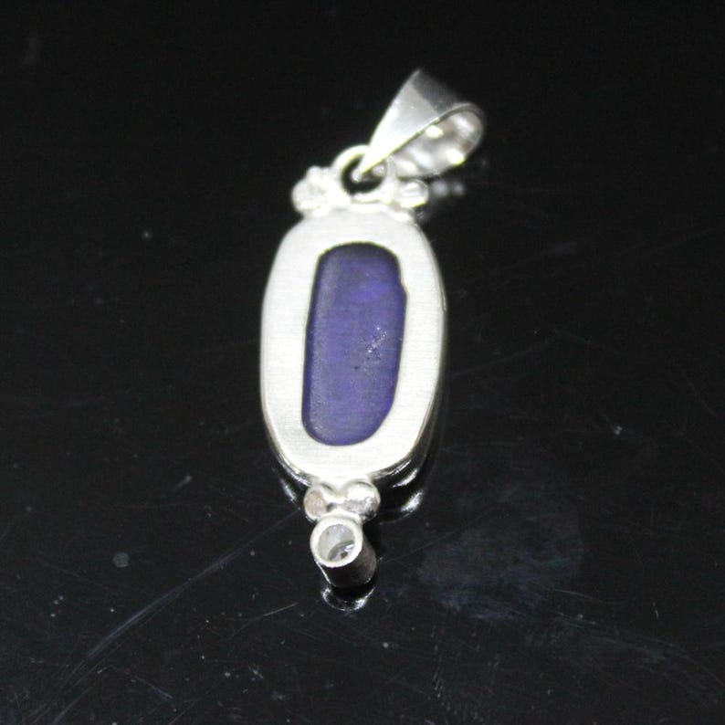 May include: A silver pendant with an oval, deep blue stone. The pendant has a bail at the top for a chain and a small decorative element at the bottom. The stone is set in a silver bezel.