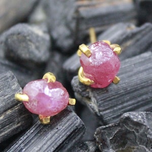 May include: A pair of ruby stud earrings. Each earring features a raw, pinkish-red ruby stone held in a gold-colored setting. The earrings are displayed on a dark, textured surface.