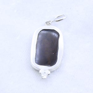 May include: A rectangular pendant with a dark brown center stone set in a silver-colored metal frame. The pendant has a loop at the top for a chain and a decorative three-petal design at the bottom.