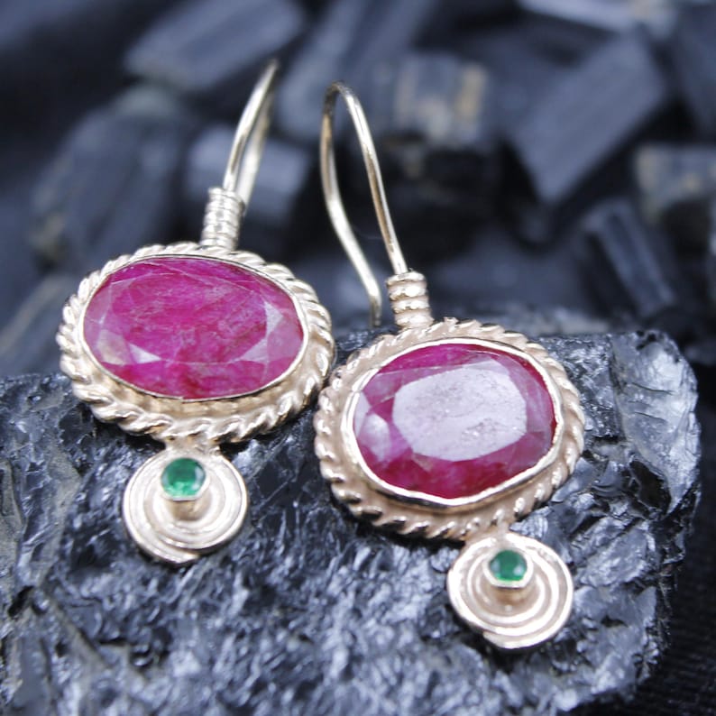 May include: A pair of gold-colored earrings featuring oval-shaped, deep red gemstones. Each earring has a smaller green gemstone below the larger stone, set in a gold-toned frame with a twisted rope design.