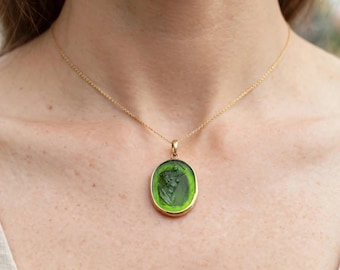 Puede incluir: Un collar de oro con un colgante ovalado que presenta un centro de vidrio verde translúcido. La pieza de vidrio muestra un relieve tallado de una figura de perfil. El colgante cuelga de una delicada cadena de oro sobre la clavícula.