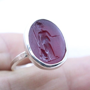 May include: A silver ring with a red carnelian stone featuring an engraved image of a woman holding a staff.