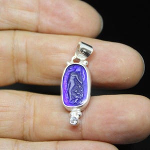 May include: A silver pendant with a rectangular, purple-hued stone featuring a carved figure. The pendant has a small loop at the top and a small, clear stone at the bottom. The pendant is held in a hand.