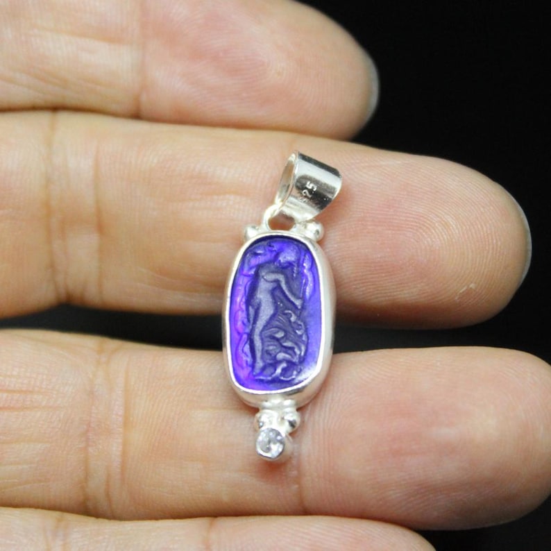 May include: A silver pendant with a rectangular, purple-hued stone featuring a carved figure. The pendant has a small loop at the top and a small, clear stone at the bottom. The pendant is held in a hand.