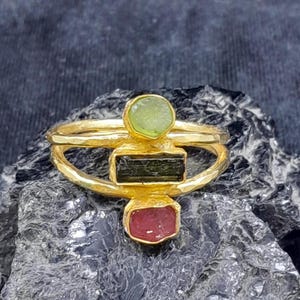 May include: A gold-colored ring with three gemstones. The ring features a round, light green gemstone at the top, a rectangular, dark green gemstone in the middle, and a red gemstone at the bottom. The ring is set on a dark, textured surface.
