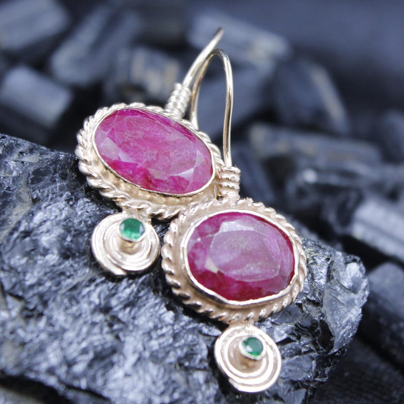 May include: Gold-toned earrings featuring large, oval-cut ruby gemstones. Each earring has a textured, rope-like border around the ruby and a small, round emerald accent. The earrings are displayed on a dark, textured surface.
