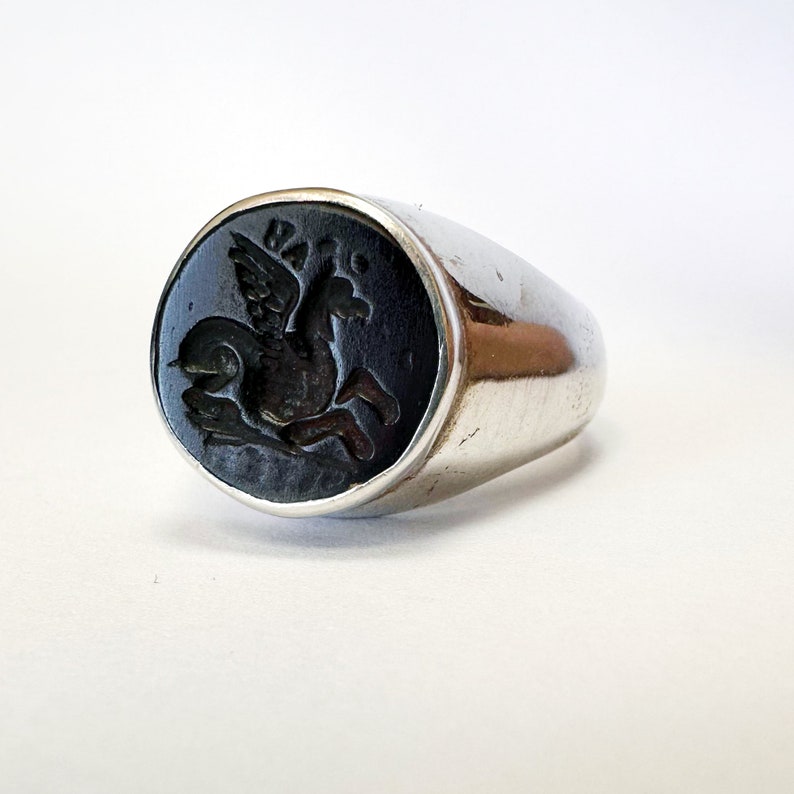 May include: A silver ring with a black onyx stone featuring a carved image of a winged horse.