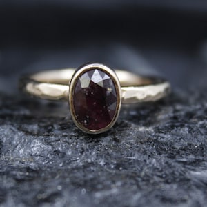 Può includere: Un anello con una pietra preziosa sfaccettata di colore rosso scuro di forma ovale incastonata in una lunetta. La fascia è realizzata in metallo di colore chiaro e presenta una finitura martellata e testurizzata sulla superficie.