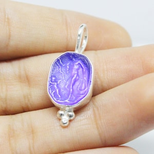 Puede incluir: Un colgante de plata con un camafeo de vidrio morado que presenta un diseño detallado. El colgante tiene un aro y tres pequeñas cuentas en la parte inferior.
