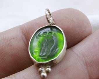 Puede incluir: Un colgante de plata con un camafeo de vidrio verde con un diseño tallado. El colgante tiene un aro y tres pequeñas bolas decorativas en la parte inferior.
