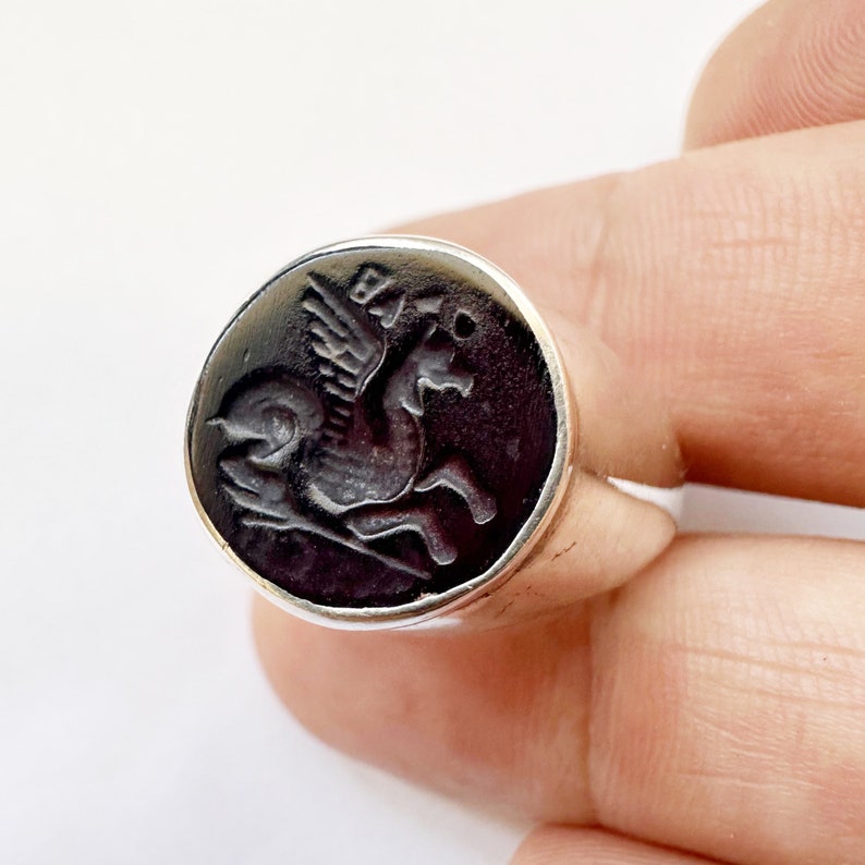 May include: A silver ring with a black onyx stone featuring a detailed engraved image of a winged horse.