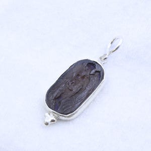 May include: A silver pendant with a dark, rectangular stone featuring a carved figure. The stone is set in a silver bezel and has a small loop at the top for a chain. The bottom of the pendant has a decorative silver element.