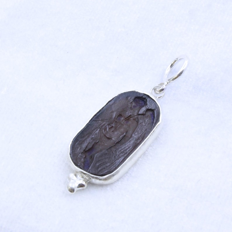 May include: A silver pendant with a dark, rectangular stone featuring a carved figure. The stone is set in a silver bezel and has a small loop at the top for a chain. The bottom of the pendant has a decorative silver element.