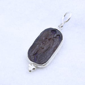 May include: A dark brown pendant with a silver-colored frame and bail. The pendant features a carved design of a figure. The bottom of the pendant has a decorative silver-colored element.