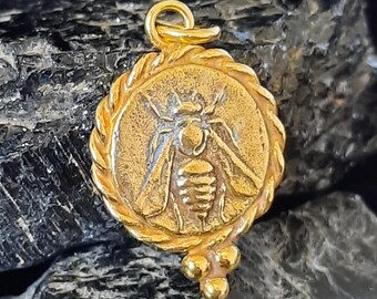 May include: A gold-toned pendant featuring a detailed bee design. The bee is surrounded by a rope-like border and the pendant has three small gold beads hanging from the bottom.