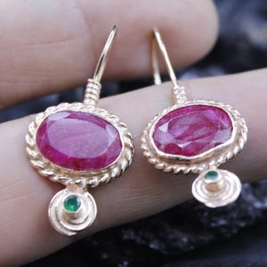 May include: Gold-toned earrings featuring oval, deep red gemstones. The gemstones are set within a twisted rope-like border. Each earring has a small, round green gemstone at the bottom. The earrings are designed with a hook closure.