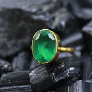 May include: A gold ring featuring a large, oval-cut emerald gemstone. The emerald is a vibrant green color and is set in a gold band. The ring is displayed on a dark, textured surface.
