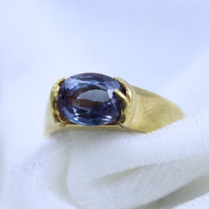 Può includere: Un anello d'oro con una grande pietra preziosa ovale di colore viola incastonata al centro. L'anello ha una fascia semplice e la pietra preziosa è circondata da una delicata montatura in oro.