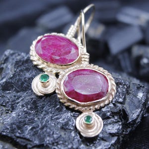 May include: Gold-toned earrings featuring large, oval-cut ruby gemstones. Each earring has a smaller, round emerald accent and a decorative rope-like border. The earrings are set against a dark, textured background.