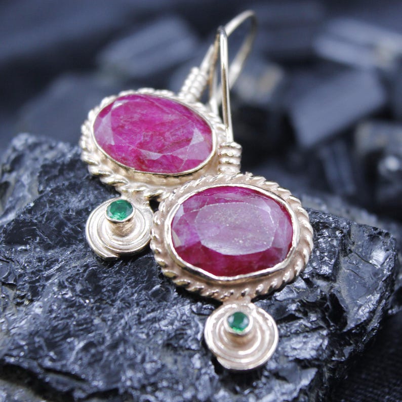 May include: Gold-toned earrings featuring large, oval-cut ruby gemstones. Each earring has a smaller, round emerald accent and a decorative rope-like border. The earrings are set against a dark, textured background.