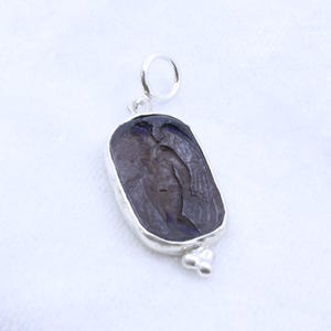 May include: A silver pendant with a dark, carved stone featuring a figure with wings. The stone is set in a silver bezel with a small loop at the top for a chain and a decorative element at the bottom.