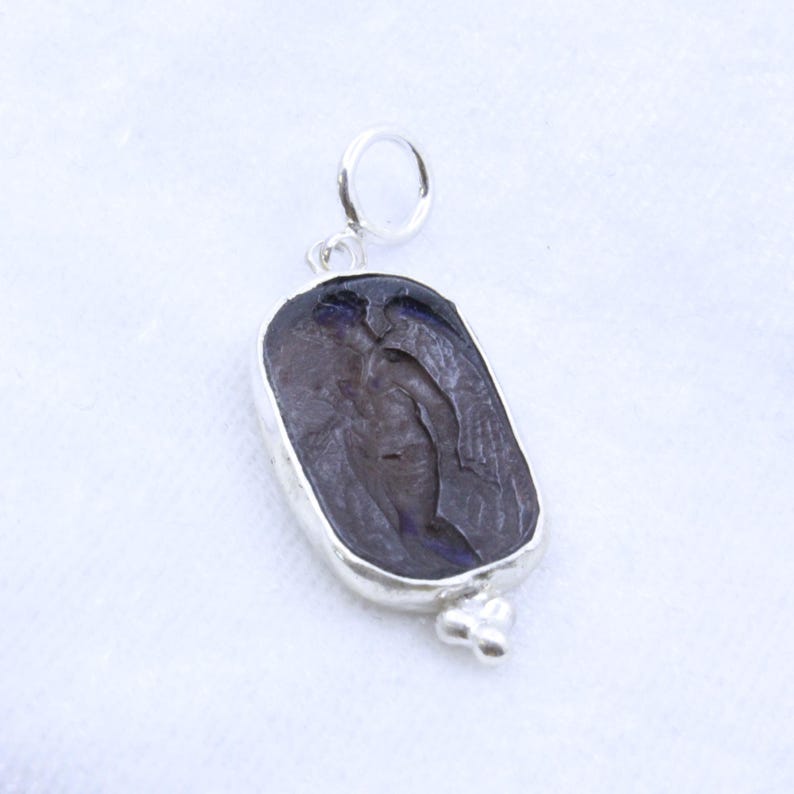 May include: A silver pendant with a dark, carved stone featuring a figure with wings. The stone is set in a silver bezel with a small loop at the top for a chain and a decorative element at the bottom.
