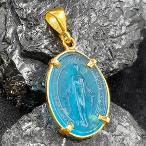Puede incluir: Pendiente de oro con un &oacute;valo de vidrio azul que representa a la Virgen Mar&iacute;a. El &oacute;valo est&aacute; rodeado por un marco dorado con la inscripci&oacute;n "Our Lady of Lourdes" en ingl&eacute;s.