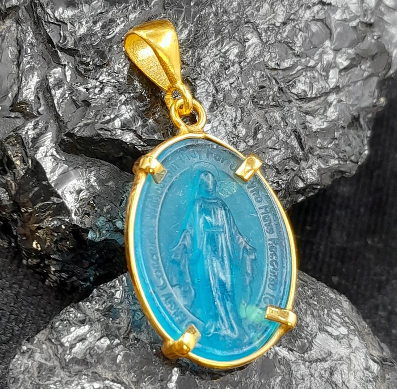 Puede incluir: Pendiente de oro con un &oacute;valo de vidrio azul que representa a la Virgen Mar&iacute;a. El &oacute;valo est&aacute; rodeado por un marco dorado con la inscripci&oacute;n "Our Lady of Lourdes" en ingl&eacute;s.