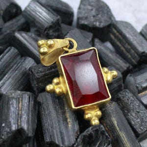 Könnte beinhalten: Ein goldener Anhänger mit einem rechteckigen, tiefroten Edelstein. Der Anhänger hat eine dekorative Goldfassung mit kleinen, runden Akzenten. Der Hintergrund besteht aus schwarzen, rau strukturierten Steinen.